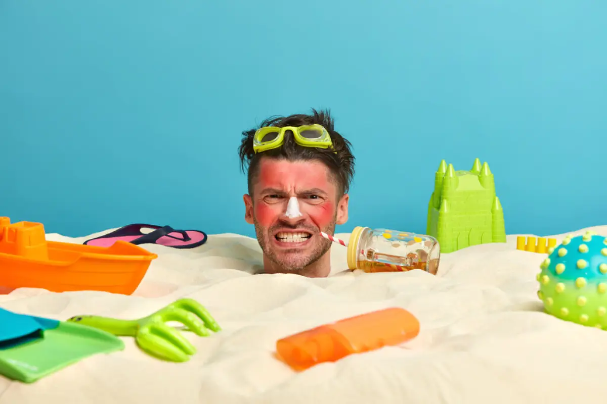 Irritaed male tourist clenches teeth from pain, being annoyed with sunburn, redness of skin, drinks cold beverage, wears goggles, poses at beach with toys, bottle of sunscreen lotion around.,  