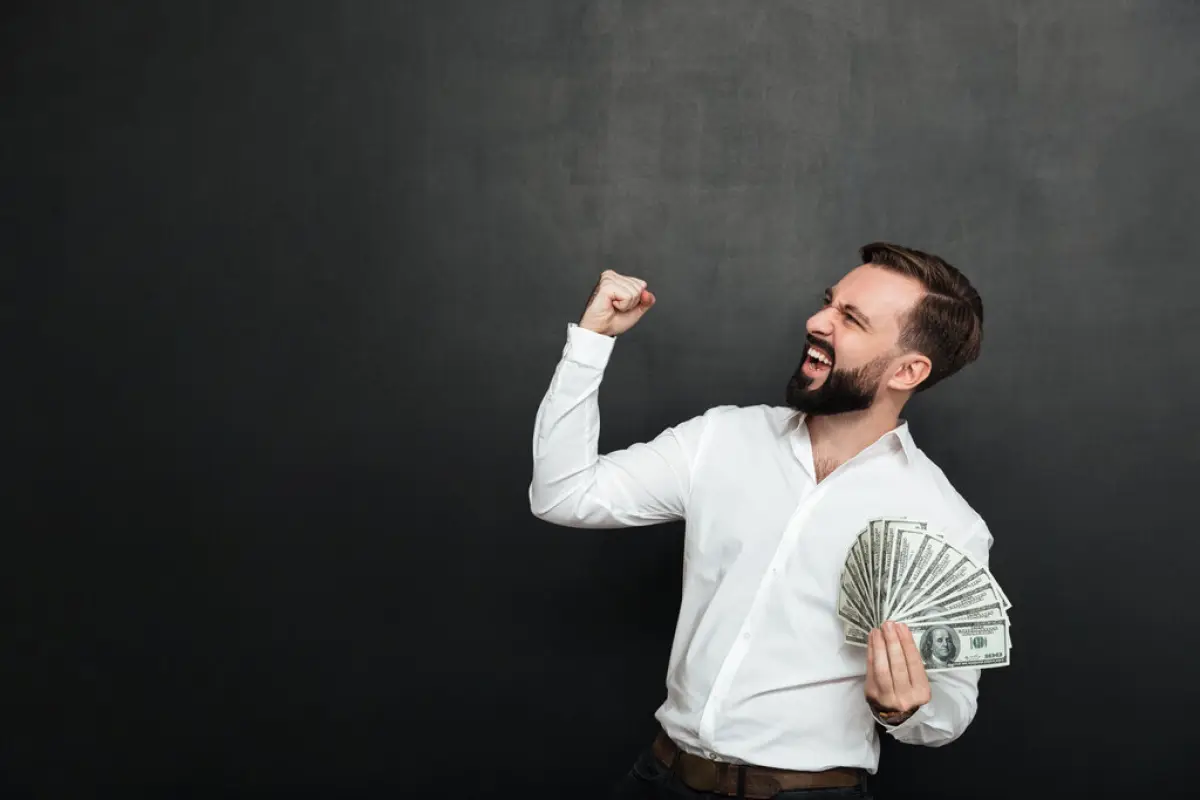 Portrait of successful guy in white shirt rejoicing like winner, Portrait of successful guy in white shirt rejoicing like winner with fan of 100 dollar bills in hand clenching fist aside over dark gray background