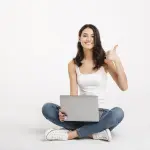 Portrait of a happy girl dressed in tank-top holding laptop, Portrait of a happy girl dressed in tank-top holding laptop while sitting on the floor and showing thumbs up gesture isolated over white background