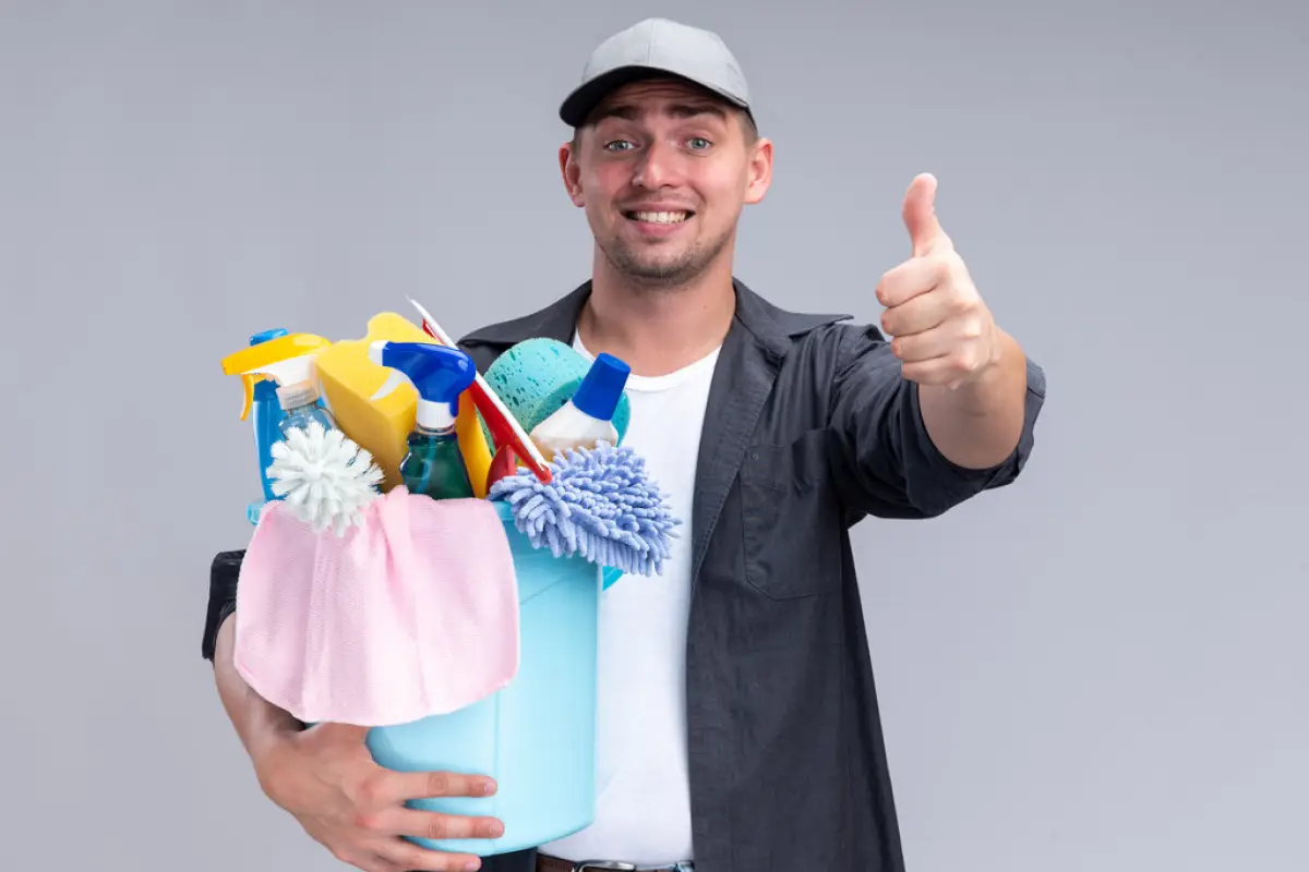 smiling young handsome cleaning guy wearing t-shirt and cap holding bucket of cleaning tools showing thumb up isolated on white background,  